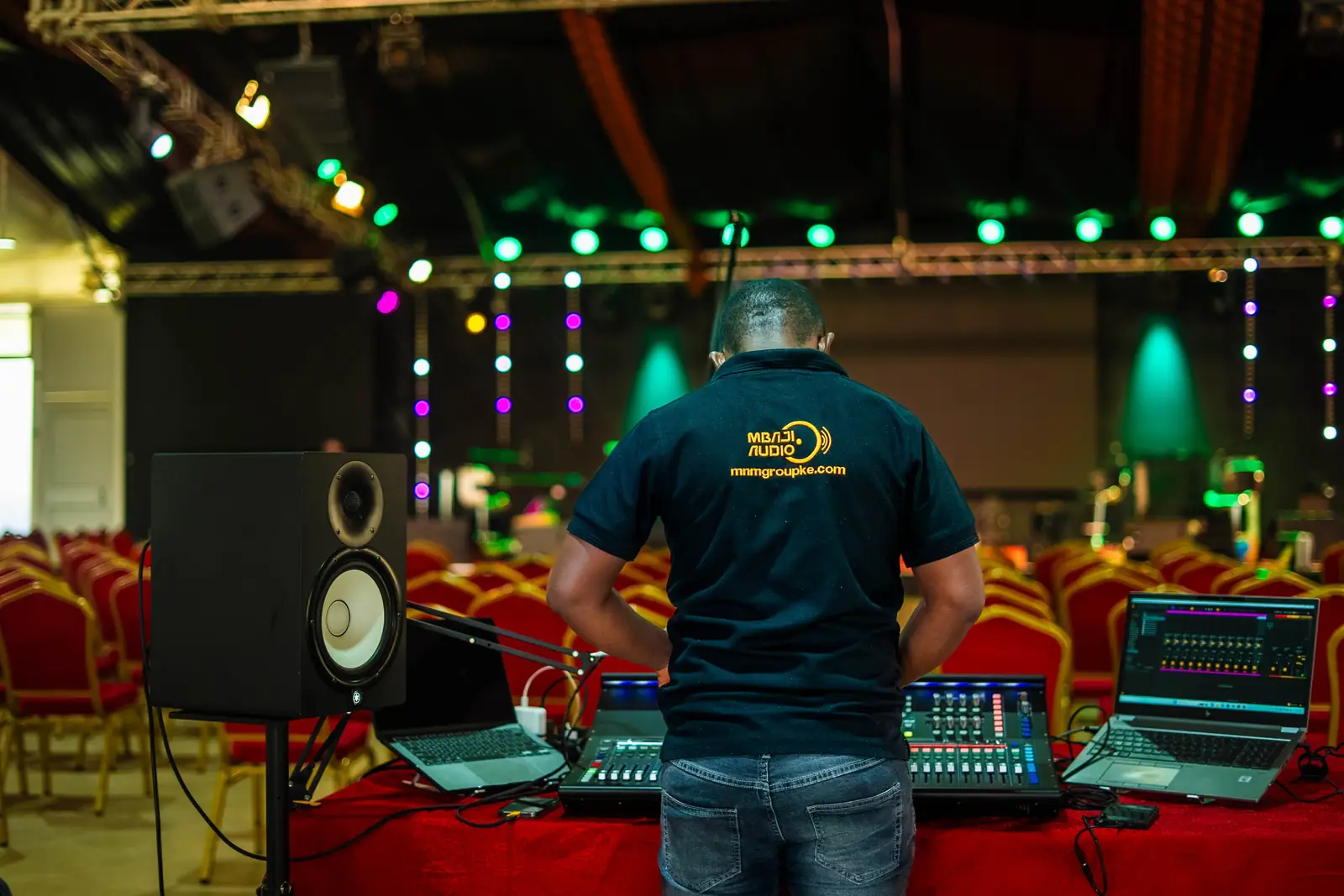 Mbaji Audio engineer at the mixing desk during an event setup, with stage lighting rigged overhead
